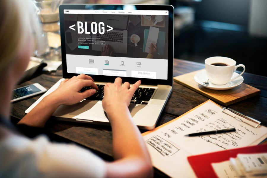 Woman seen from behind, writing a blog on a laptop in a café, with a coffee cup, pen, and papers on a wooden tabletop.