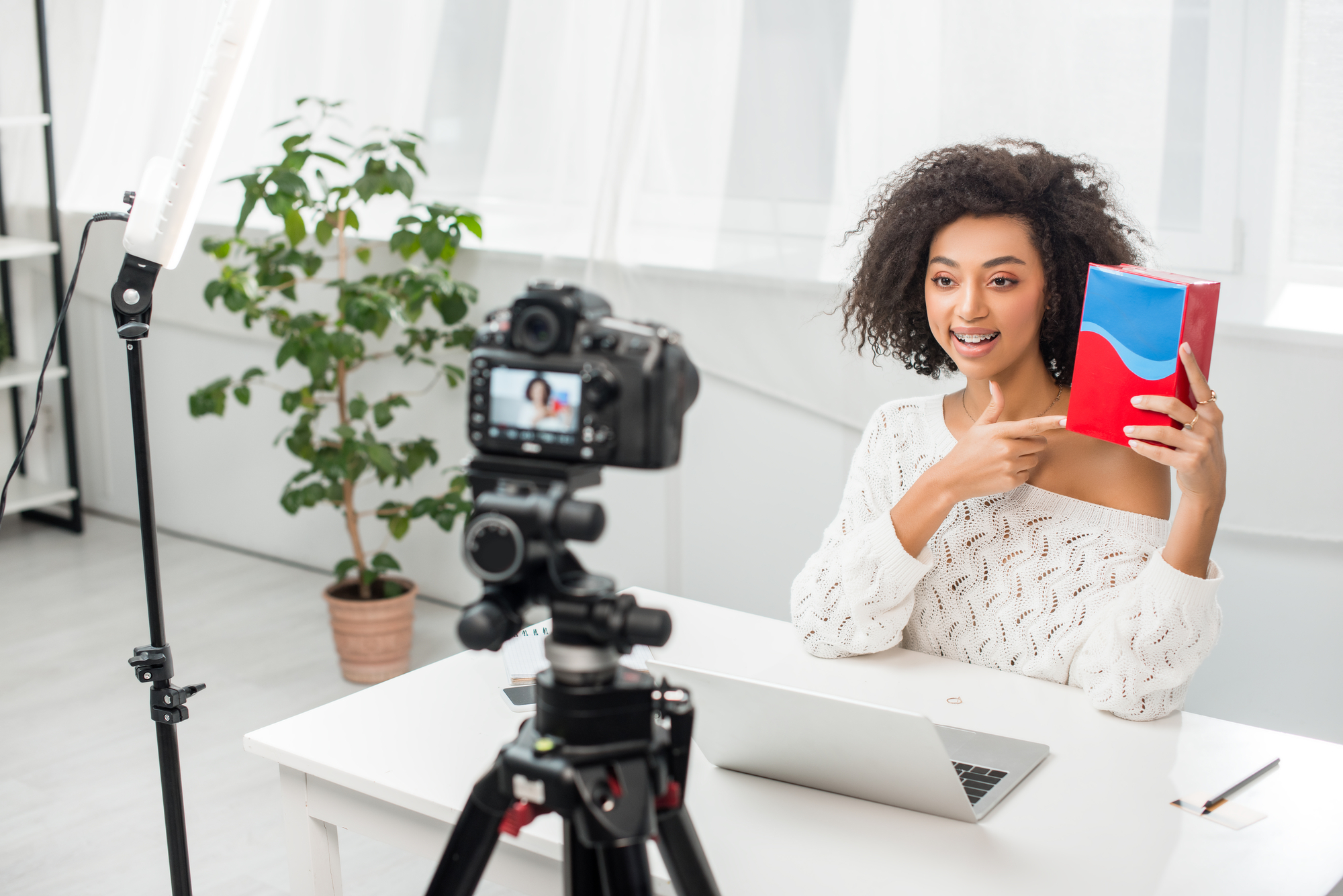 Smiling African American woman presents colorful box, with ring light on tripod and digital camera recording.