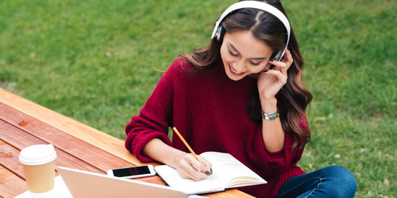 Woman in a park listening to music while writing in a notebook, with a laptop in front of her.