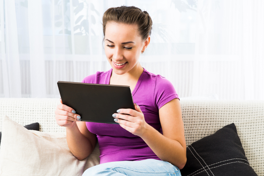 Smiling young woman with tablet on white couch in open living room.